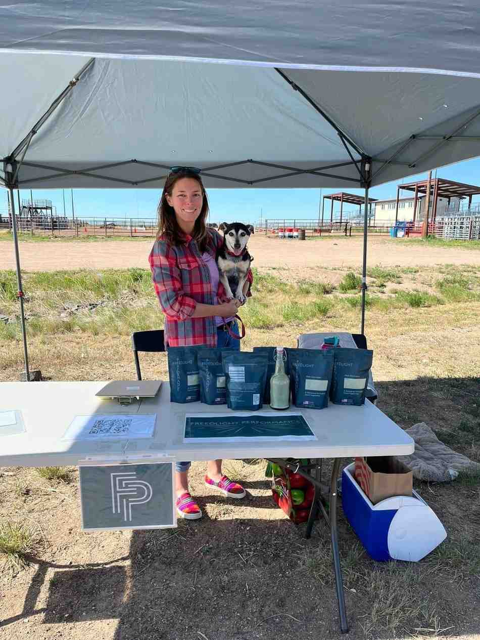 Person holding a dog and standing behind a table with products under a tent in an outdoor setting.